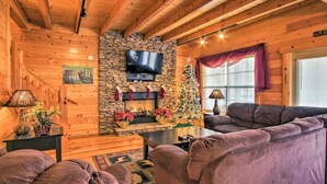 A cozy log cabin interior with a stone fireplace, a television mounted above it, and a Christmas tree. - Lazy Bear Lodge: 6BR, Theater,Spa,Pool,Game room! (Pigeon Forge)