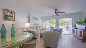 A living room with a glass table, white armchairs, a sofa, and a ceiling fan.