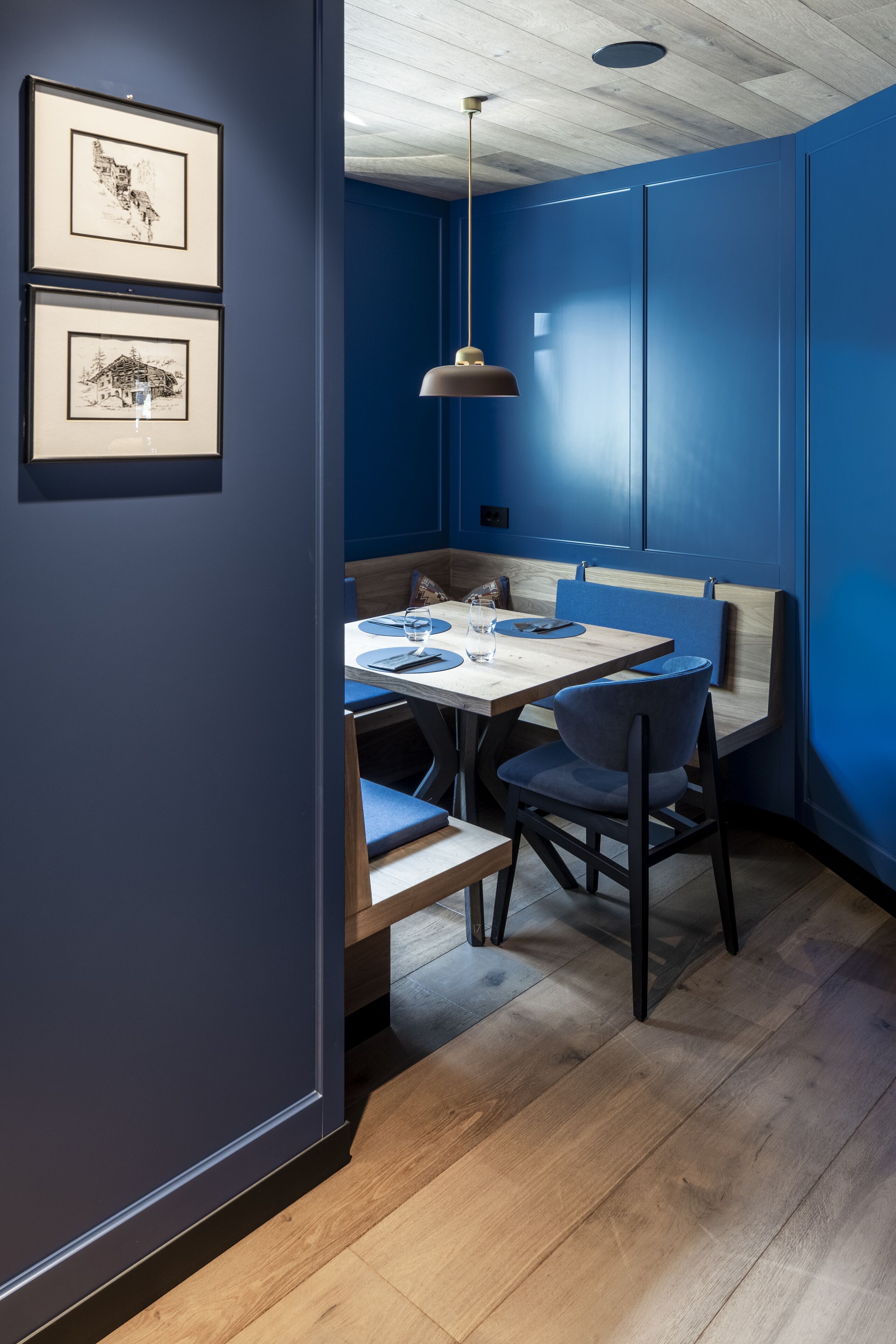 Un comedor con mesa de madera y sillas tapizadas en azul, una obra de arte en la pared y una lámpara colgante.