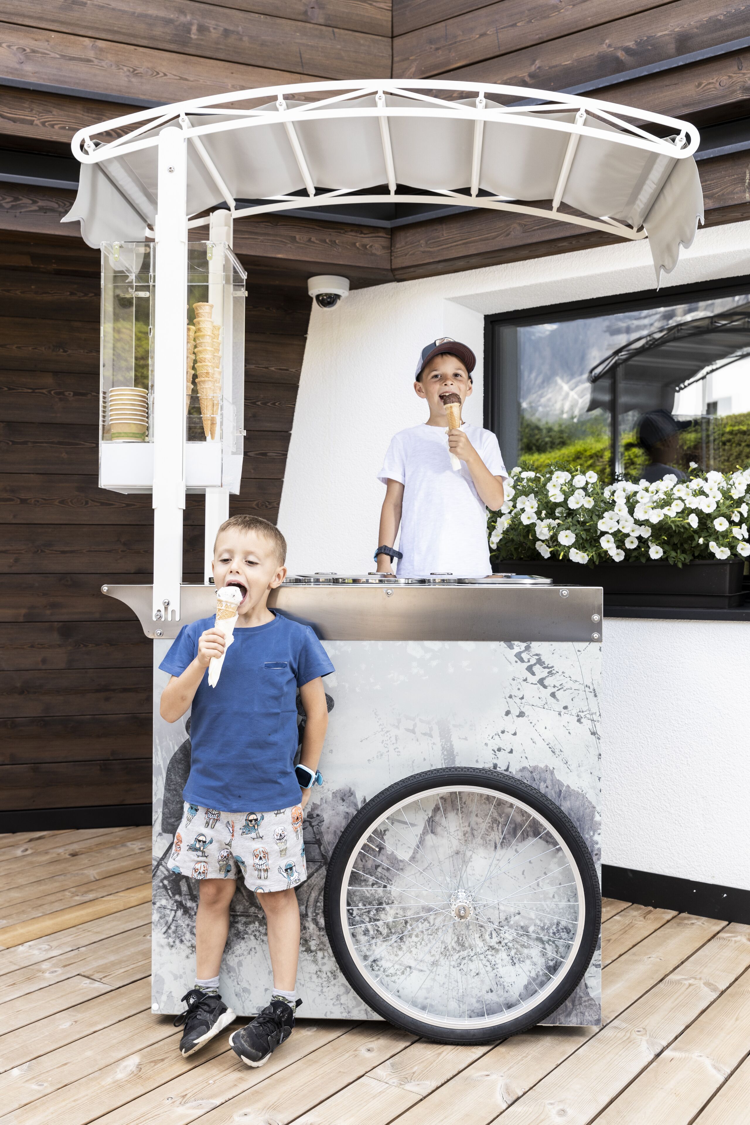 Et barn spiser is ved en udendørs kiosk med et cykelhjulsmotiv, en hvid parasol og en blomsterkasse i baggrunden.