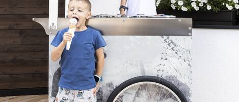 A child eating ice cream at an outdoor kiosk with a bicycle wheel design, a white umbrella, and a flower planter in the background.