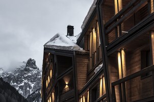 Un edificio de varios pisos con fachada de madera y amplias ventanas, un techo cubierto de nieve y montañas al fondo.