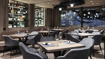 Un restaurante con varias mesas y sillas, una zona de bar con estantes llenos de botellas, y vistas a un paisaje nevado a través de amplios ventanales.