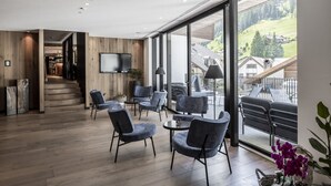 Un interior moderno con suelo de madera, sillones tapizados en gris, un televisor de pantalla plana y un balcón con vistas a un paisaje montañoso.