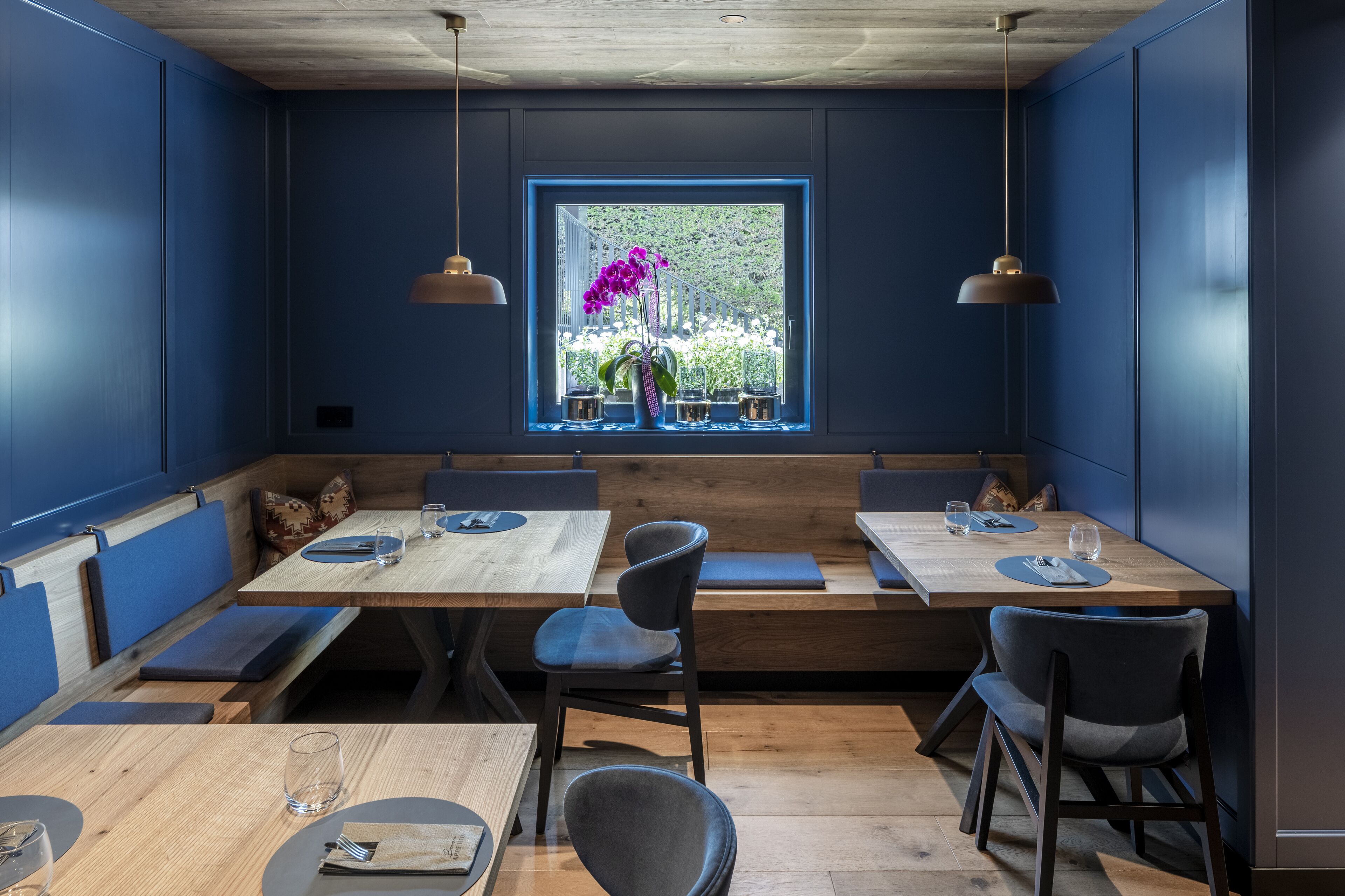 Un restaurant aux murs bleus, avec des tables et des bancs en bois. Une fenêtre offre une vue sur la verdure à l’extérieur.