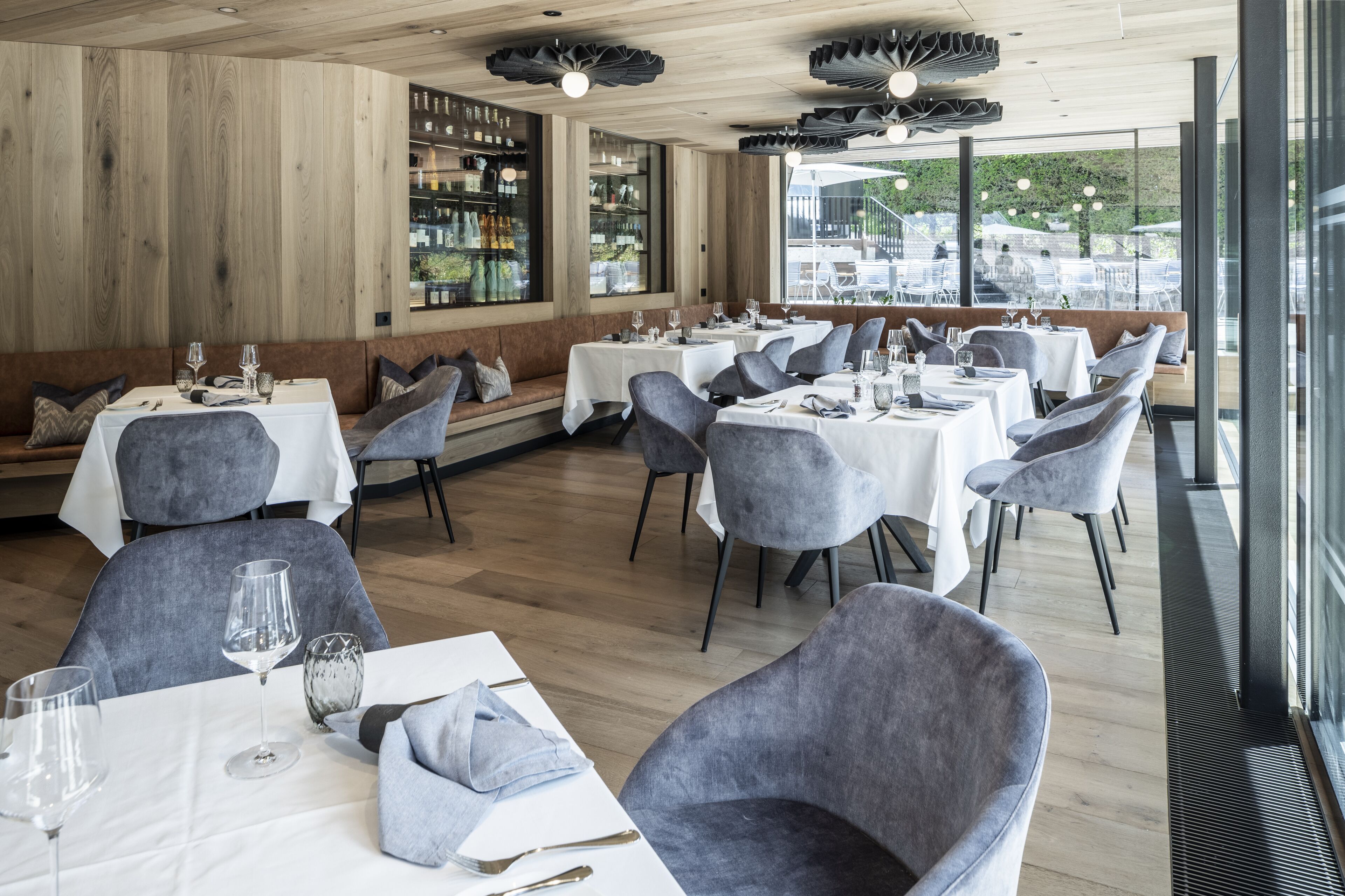 A restaurant with multiple tables and chairs, wooden walls, and large windows offering a view of the outdoors.
