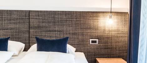 Ein ordentlich bezogenes Bett mit weißen Laken und blauen Kissen, ein kleiner Nachttisch aus Holz und eine strukturierte Wand.