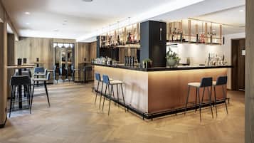 A modern bar area with a herringbone wooden floor, bar stools, and a well-lit bar counter with bottles and glasses.