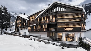 Un hotel moderno con fachada de madera y varios balcones, con imponente paisaje nevado de montañas al fondo.