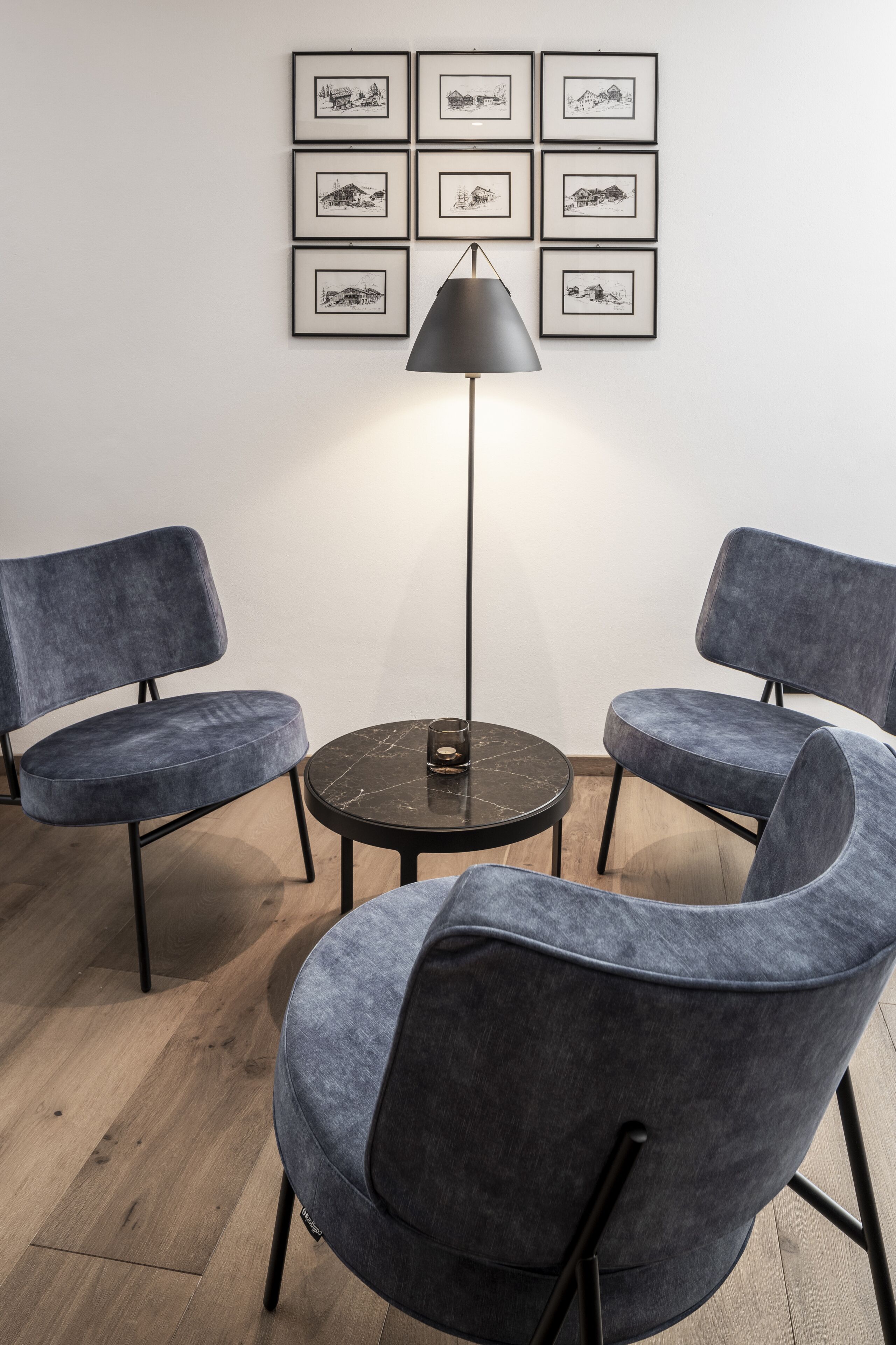 Ein Zimmer mit zwei grauen Polstersesseln um einen kleinen runden Tisch, einer schwarzen Stehlampe und sechs gerahmten schwarz-weißen Zeichnungen an der Wand.