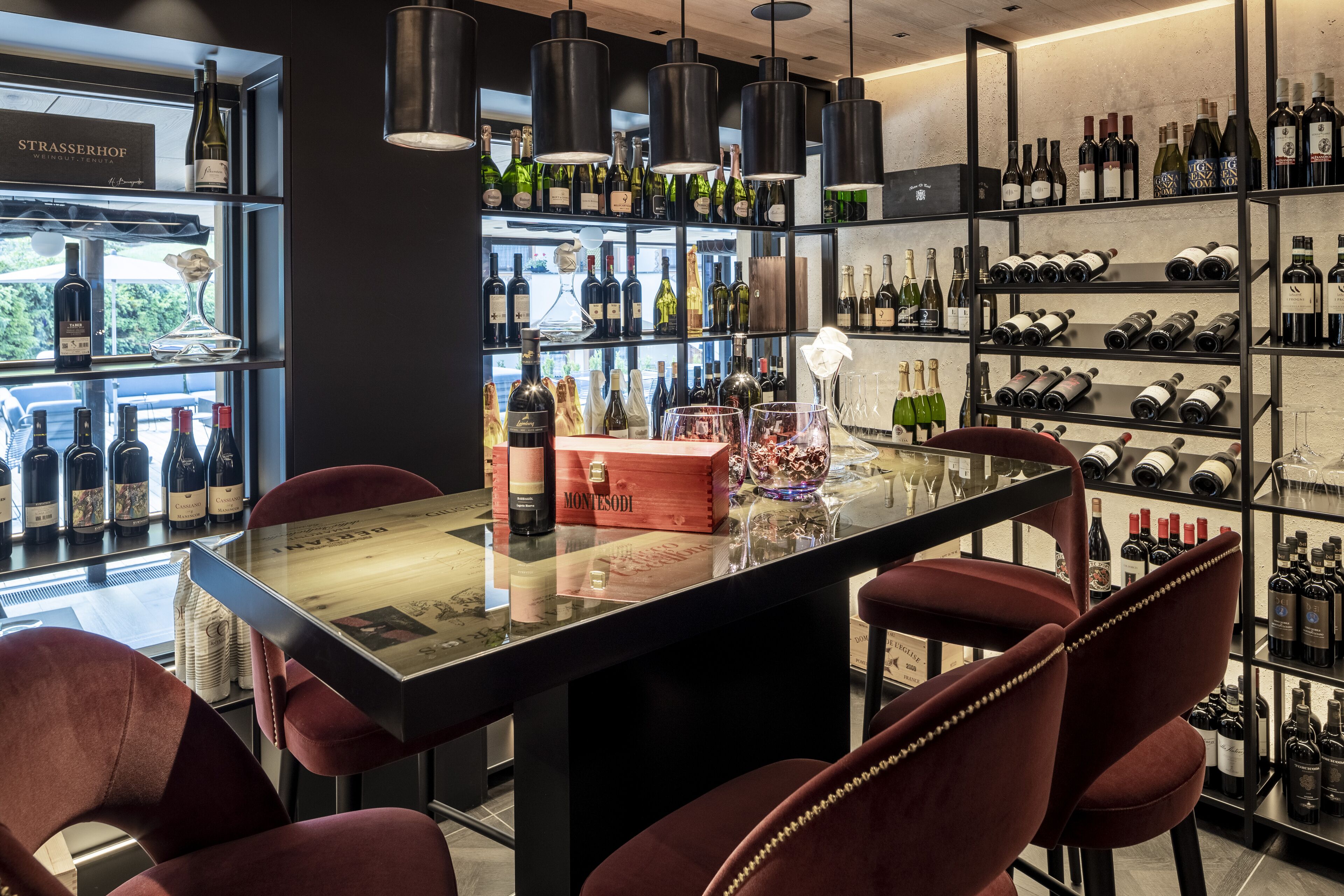 A bar area with a long counter, multiple wine bottles on shelves, and plush red chairs.