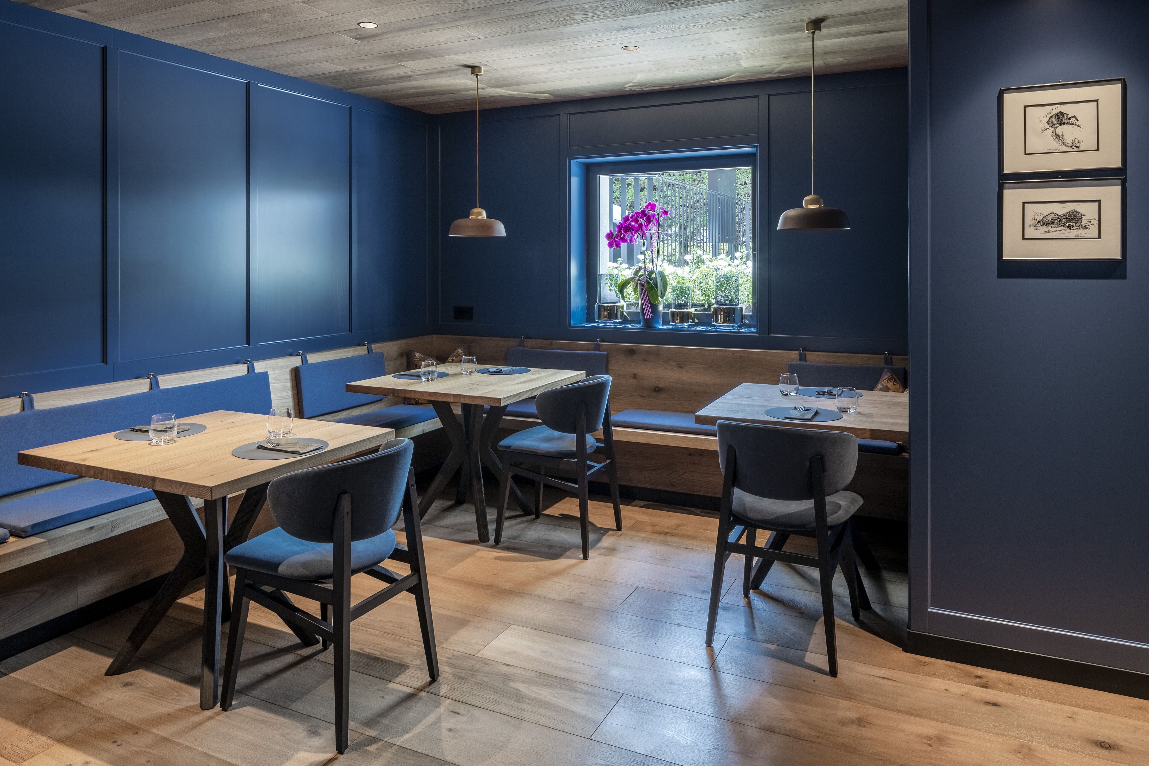 Ein Restaurant mit Holztischen und blauen Sitzbänken, gerahmten Kunstwerken an der Wand und einem Fenster mit Blick auf Grünflächen.