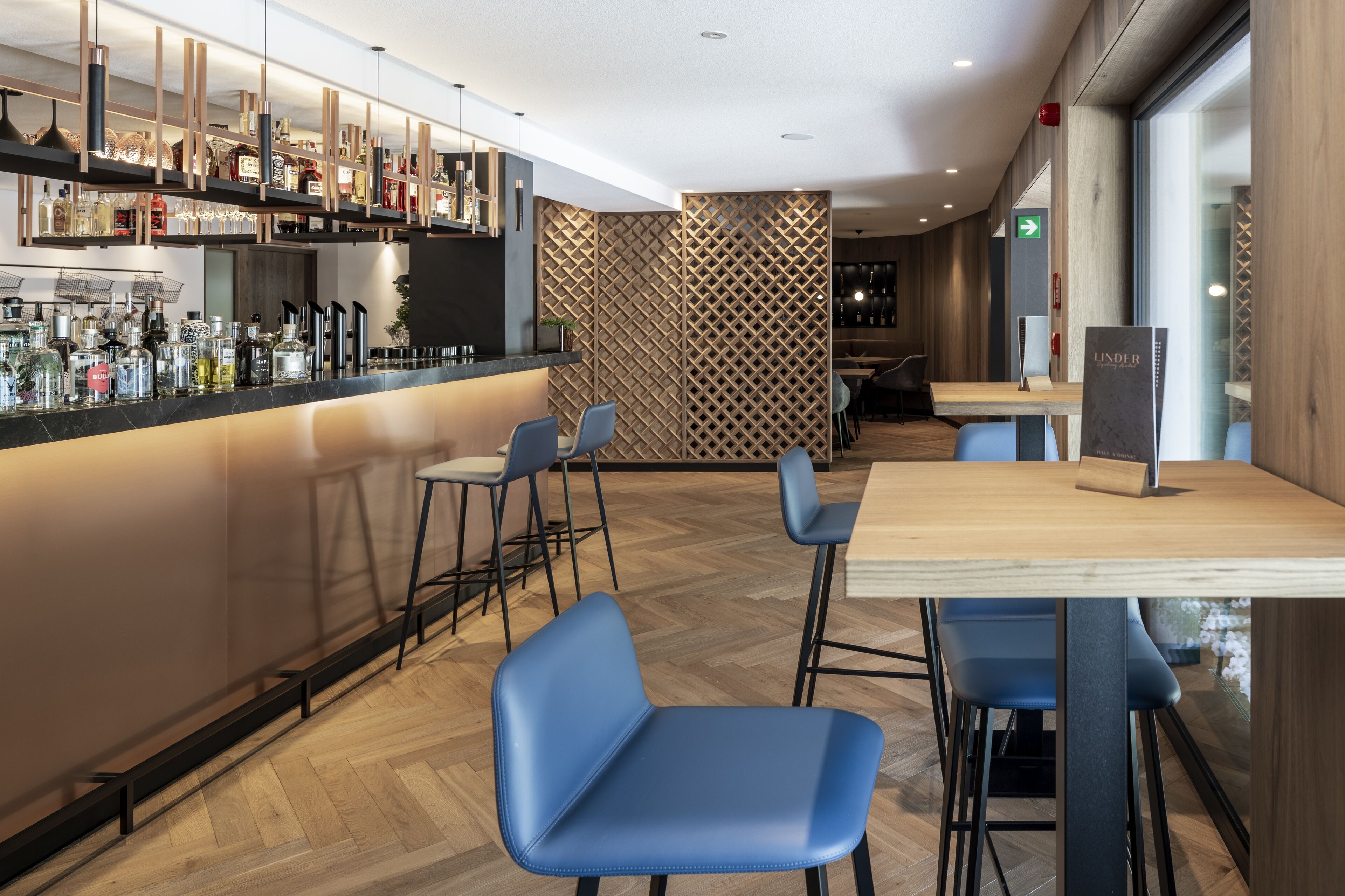 A modern bar area with a wooden floor, blue chairs, and a variety of bottles and glasses on shelves.