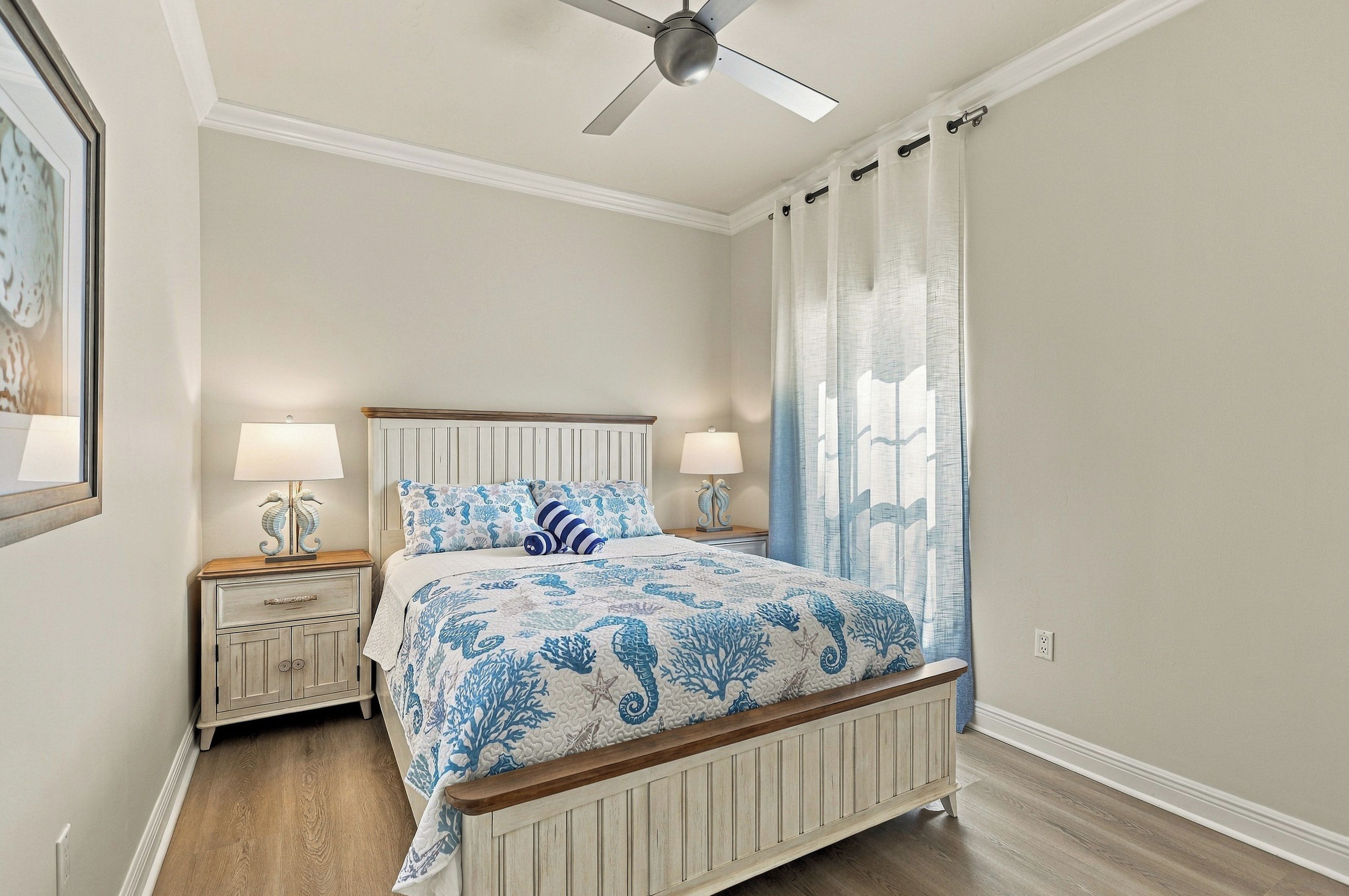 Une chambre à coucher comprenant un lit, deux tables de chevet, un ventilateur de plafond et une fenêtre avec des rideaux.