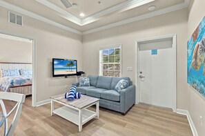 A room with a grey sofa, a television on the wall, a wooden coffee table, and a bed with a blue and white patterned bedspread. - Gulf Waters-Beach Club at Siesta Key 108D by RVA (Sarasota)