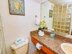 A bathroom with a toilet, sink, and mirror. There is a towel rack with towels, a flower vase, and a decorative piece on the wall. - Highly Upgraded West Maui Condo! WiFi, Modern Kitchen, Lanai–Kamaole Sands 9304 (KIHEI)