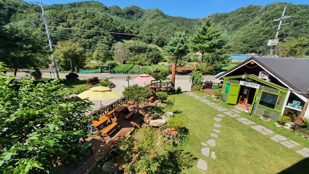 Een klein gebouw met een groen dak, met een bord in het Koreaans, omgeven door een verzorgde tuin met een pad van stenen, bankjes en een gele parasol.