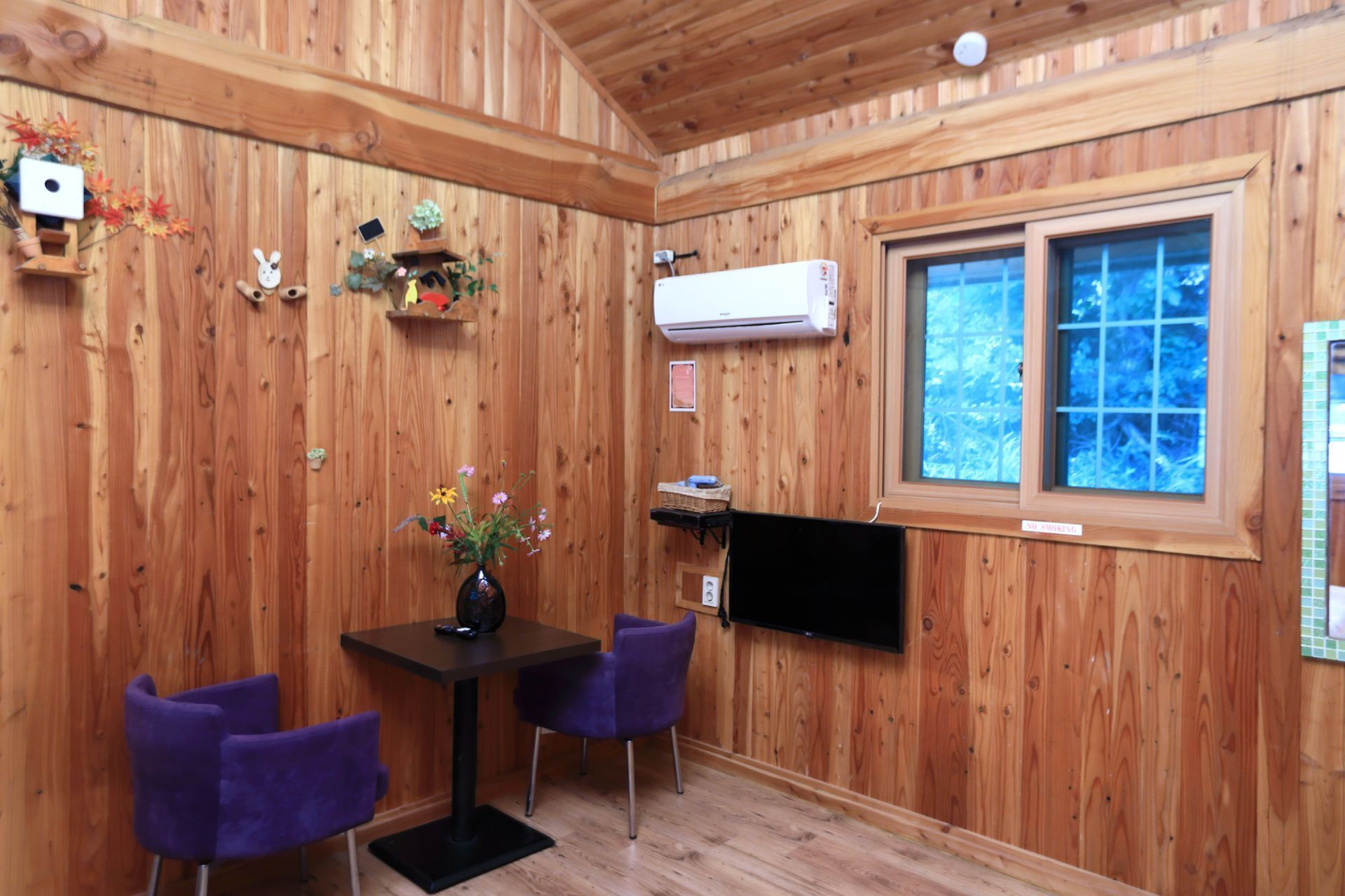 A wooden interior with a dining table and chairs, a mounted TV, and a window with a view of trees.