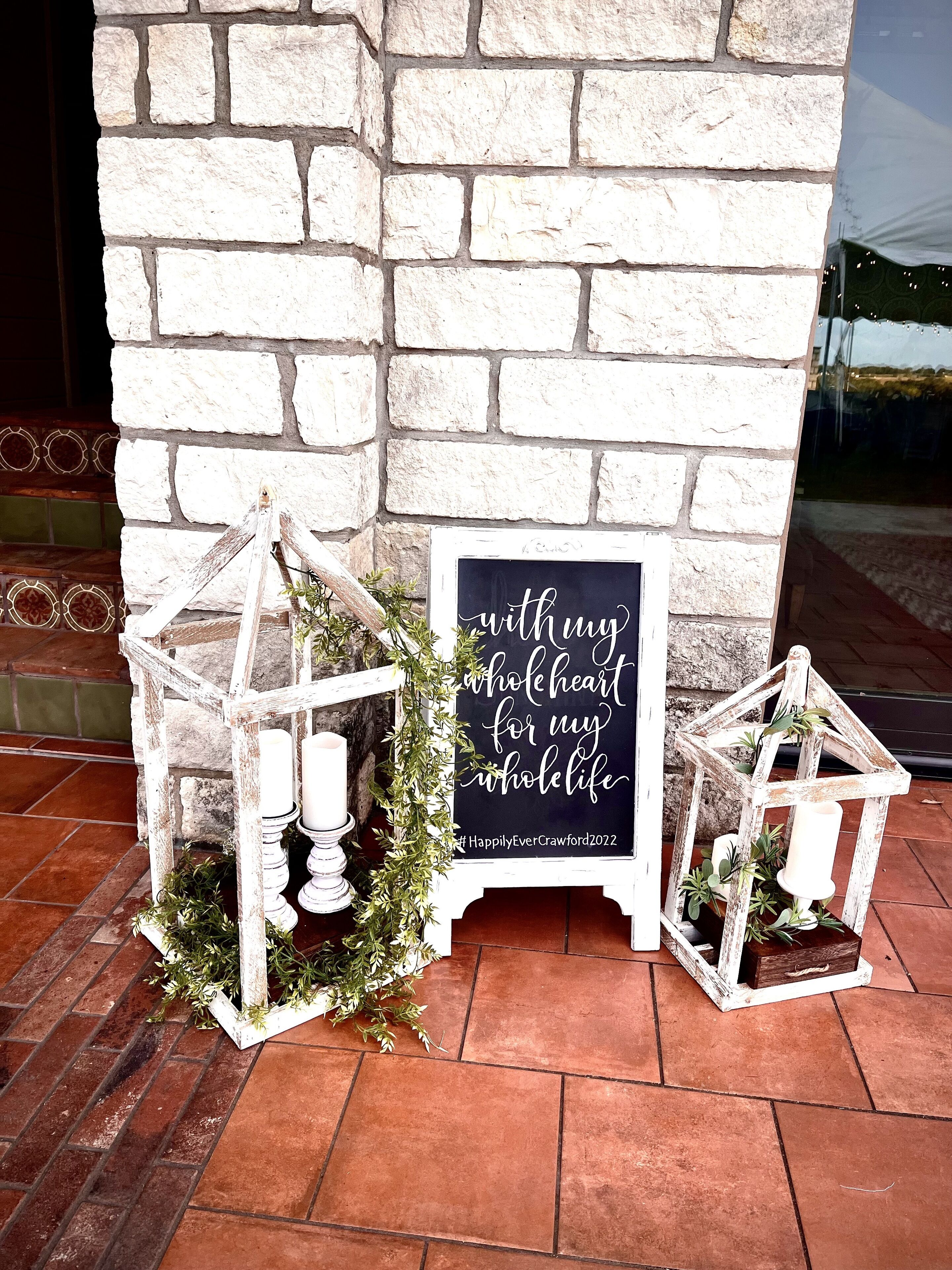 A rustic outdoor setting with a white brick wall, two white lanterns, and a sign with a heartfelt message.