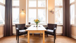 A room with a wooden table and two chairs, a vase with flowers, and a view of a building through large windows.
