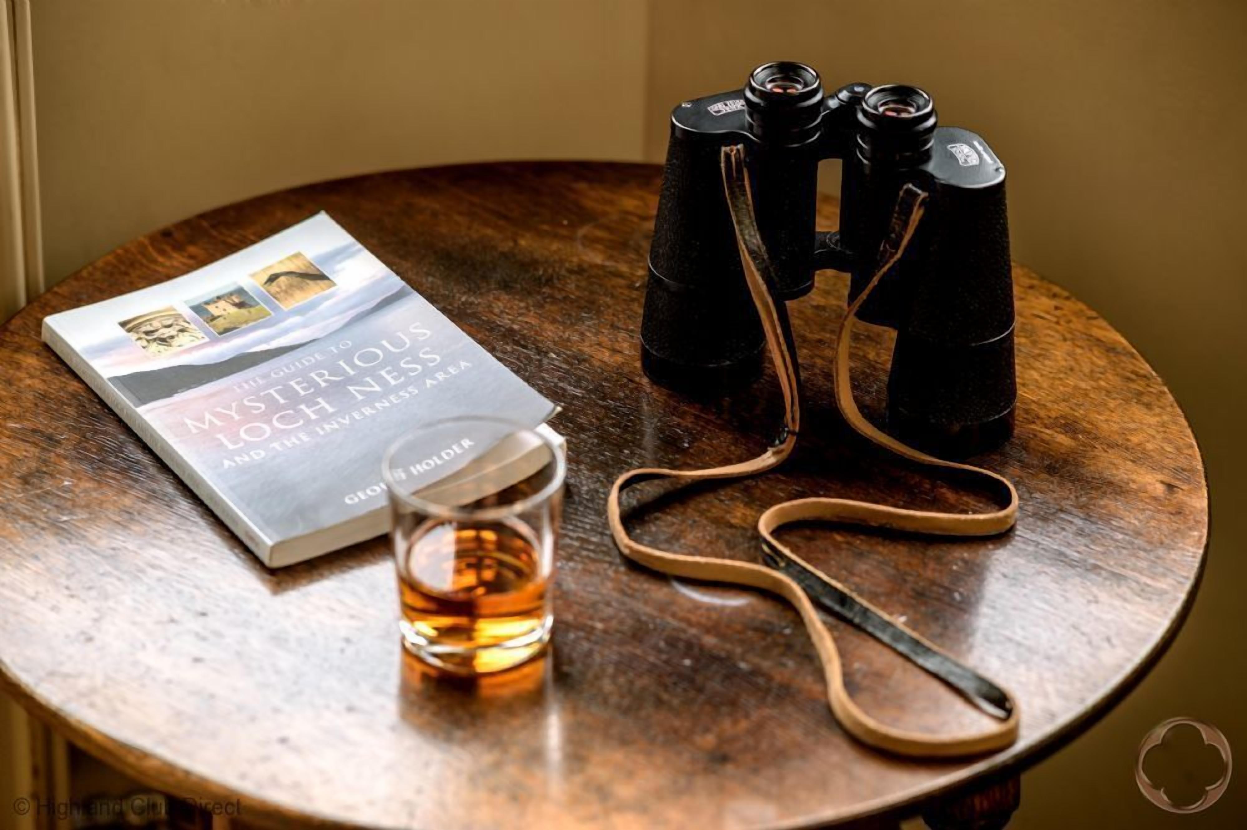 A book titled "The Guide to Mysterious Loch Ness" on a wooden table, accompanied by binoculars and a glass of amber liquid.