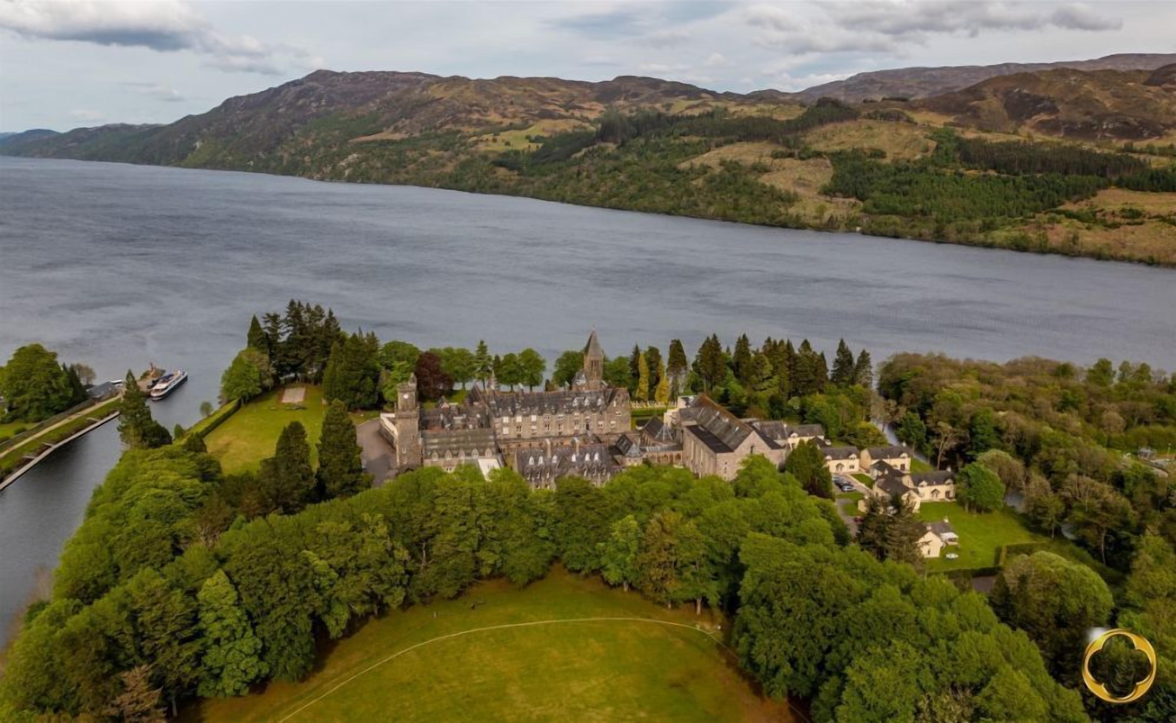 An aerial view of a historic castle with a large courtyard, surrounded by dense forest and a lake.