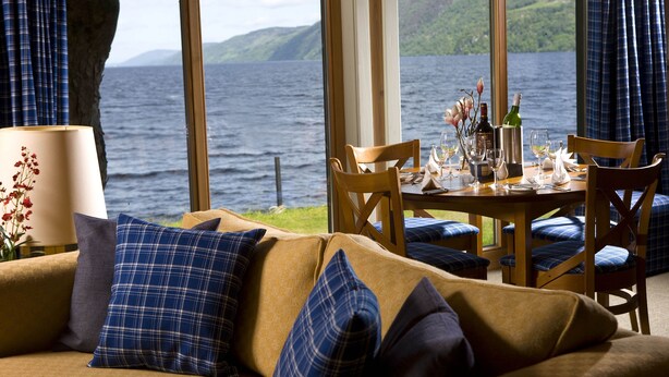 A cozy room with a large window overlooking a lake, a dining table set with a bottle, glasses, and a vase of flowers. - 9 THE MONASTERY WITH LOCH NESS VIEWS - sleeps 4 guests  in 2 bedrooms (Fort Augustus)