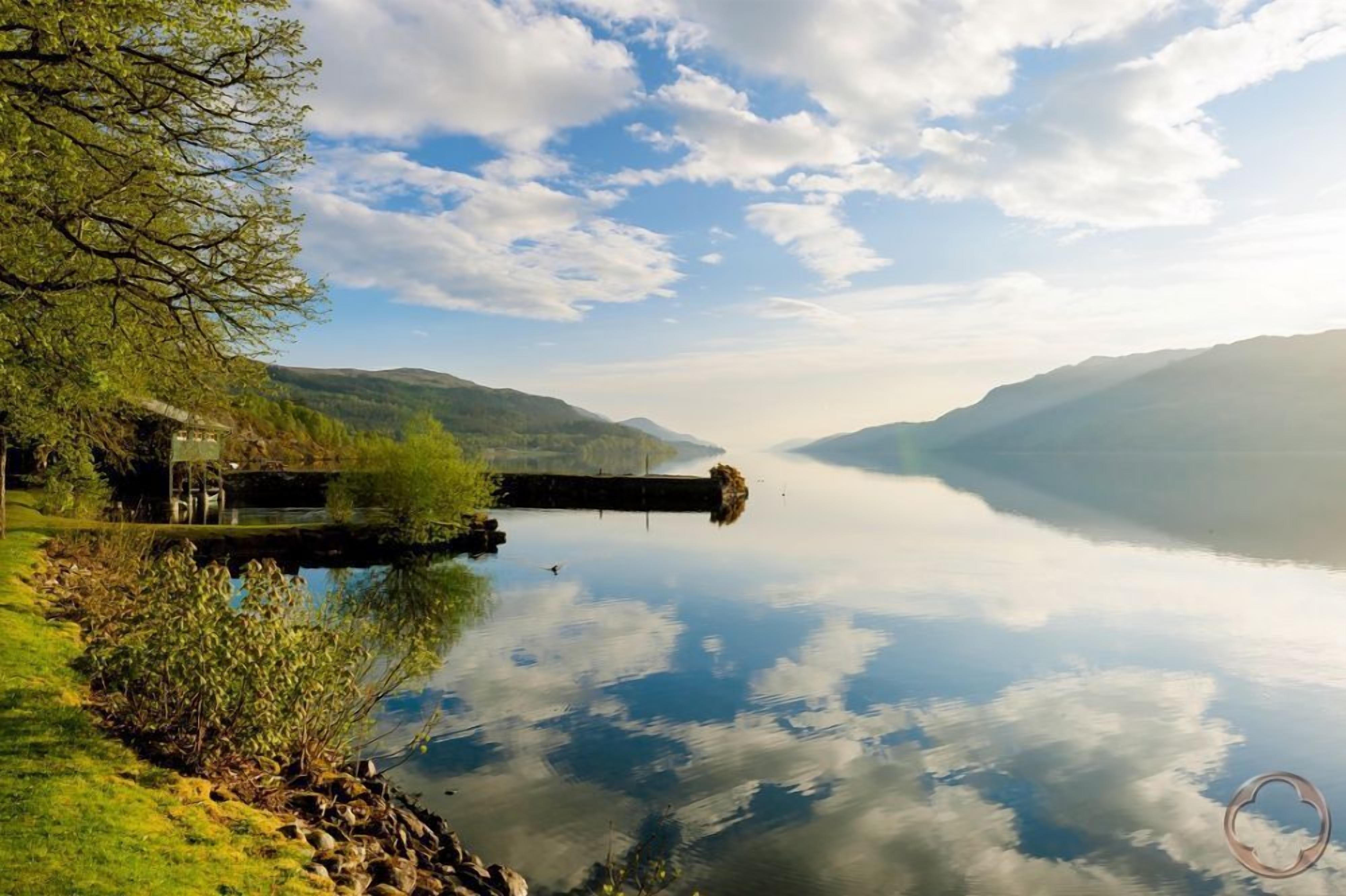 A calm lake with a dock extending into the water, surrounded by lush greenery and distant hills.