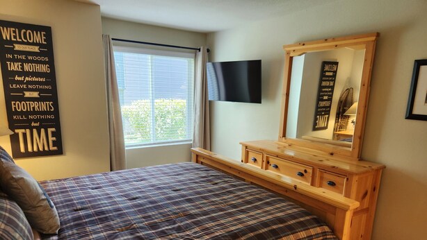 Ein Schlafzimmer mit Karo-Bettdecke, einem Holzkapfen und einem an der Wand montierten Fernseher, sowie einem Fenster mit Vorhängen.