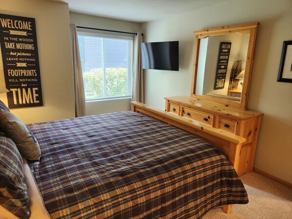 A bedroom with a plaid bedspread, a wooden dresser, a TV mounted on the wall, and a window with curtains.