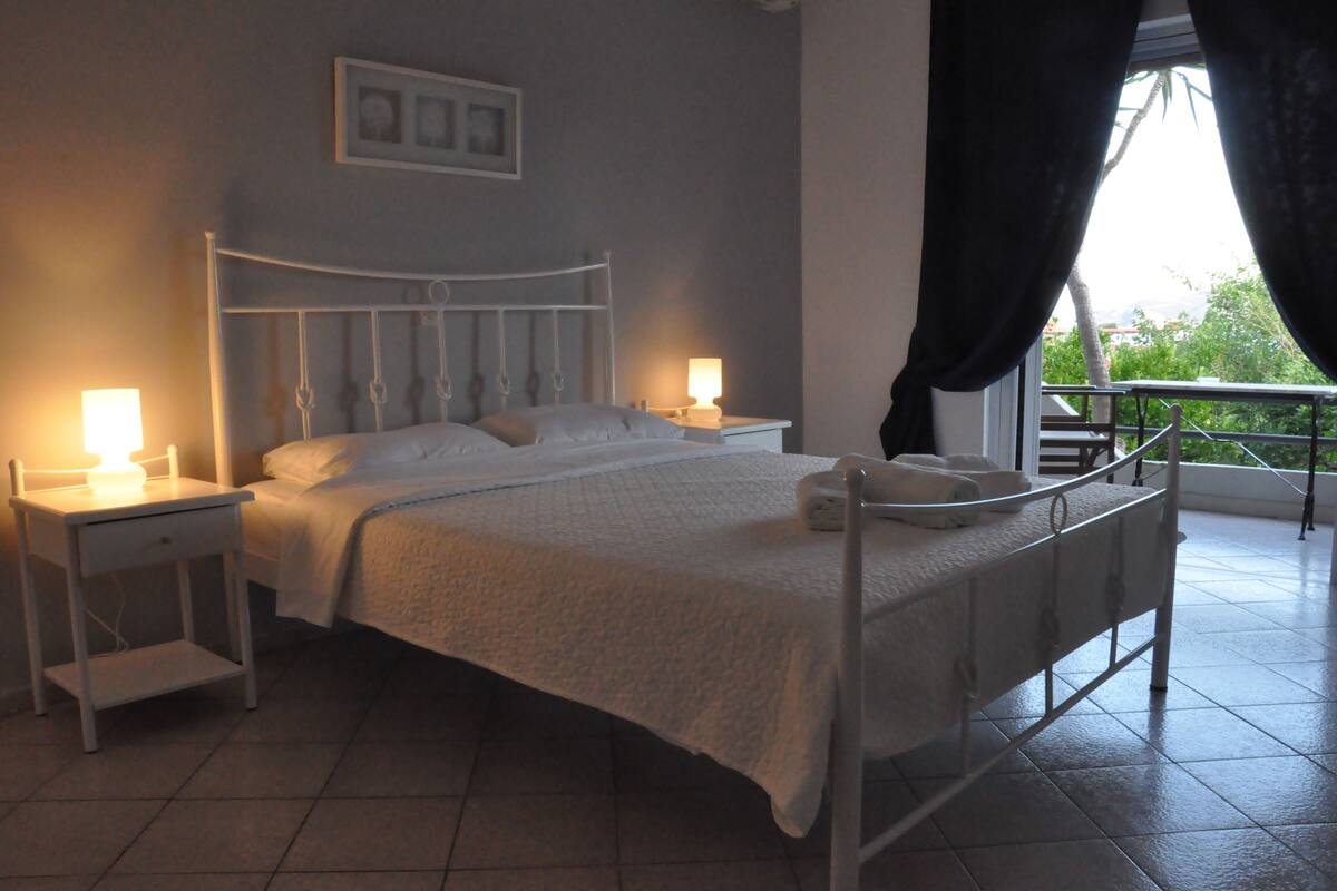 Una cama bien hecha con cabecera y piecera blancos, dos mesitas de noche con lámparas, una ventana con cortinas y la barandilla de un balcón.