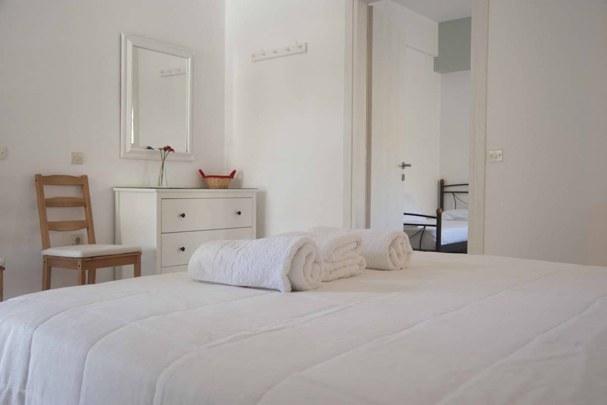 Una cama bien hecha, toallas enrolladas, una silla de madera, un tocador blanco y un espejo en la pared.