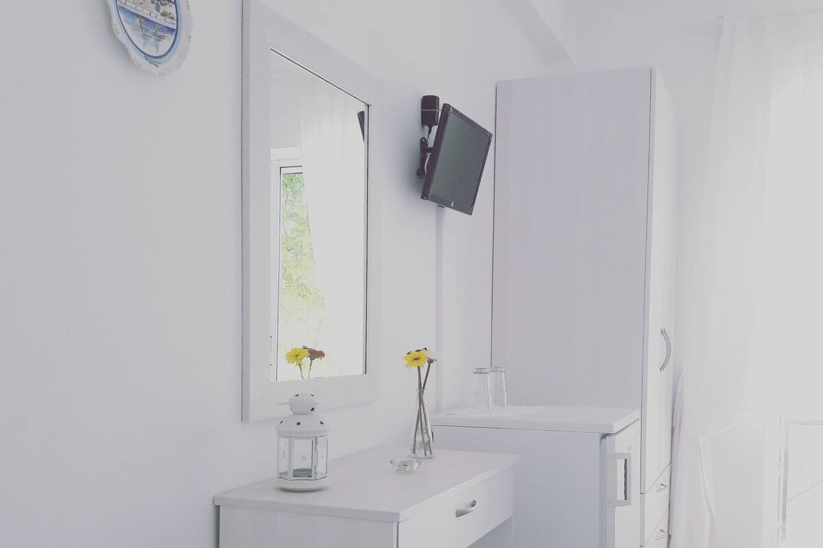 Un baño moderno con tocador blanco, un espejo, un televisor montado y un pequeño adorno en la encimera.