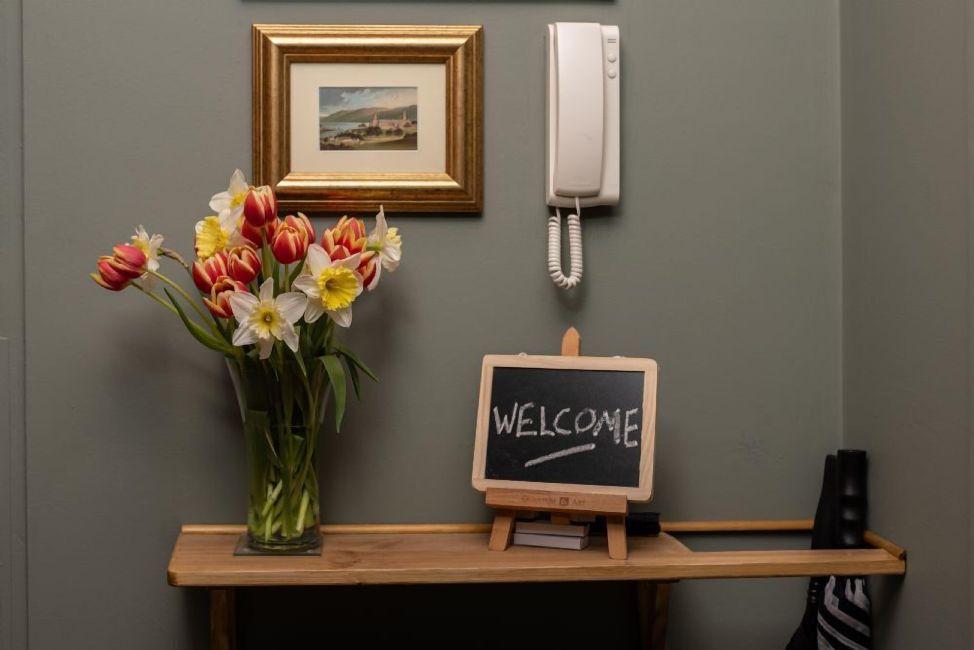 Ein Empfangstresen mit einem gerahmten Bild, einem weißen Telefon, einer Vase mit Blumen und einem Kreidetafel-Schild mit der Aufschrift 'WELCOME'.