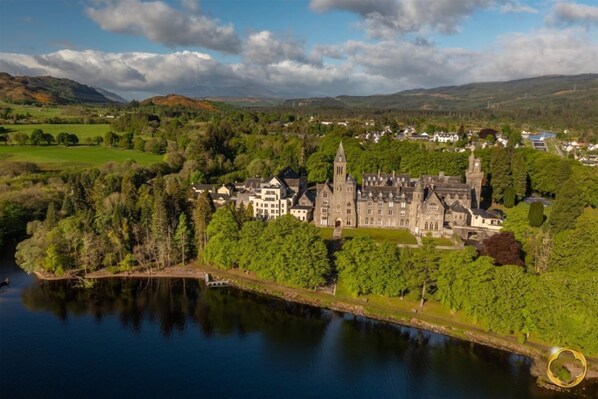 A historic castle with a large tower, situated by a lake surrounded by lush greenery and rolling hills.