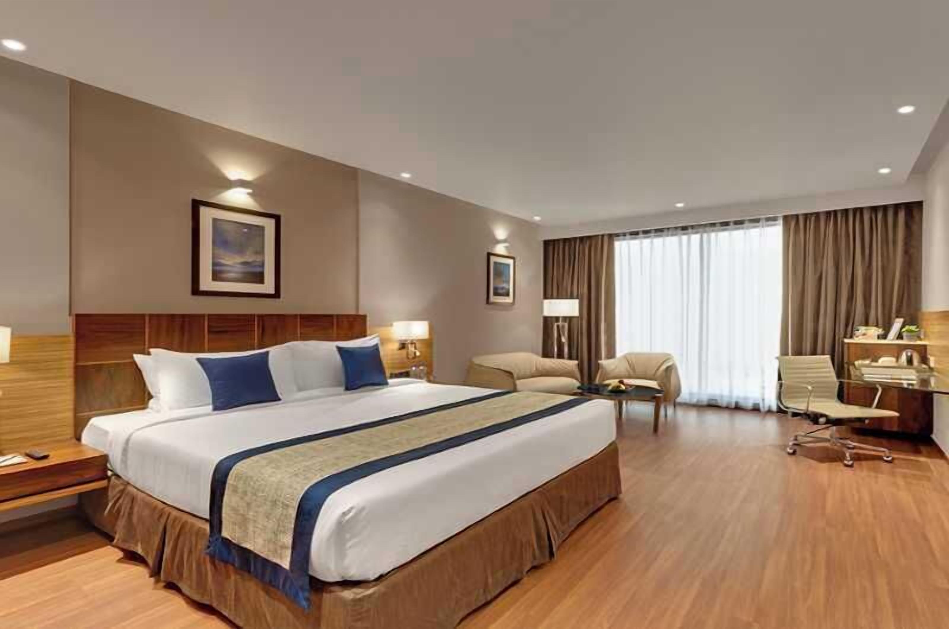 Hotelkamer met een groot bed, nachtkastjes, een bureau, een stoel en een groot raam met gordijnen.