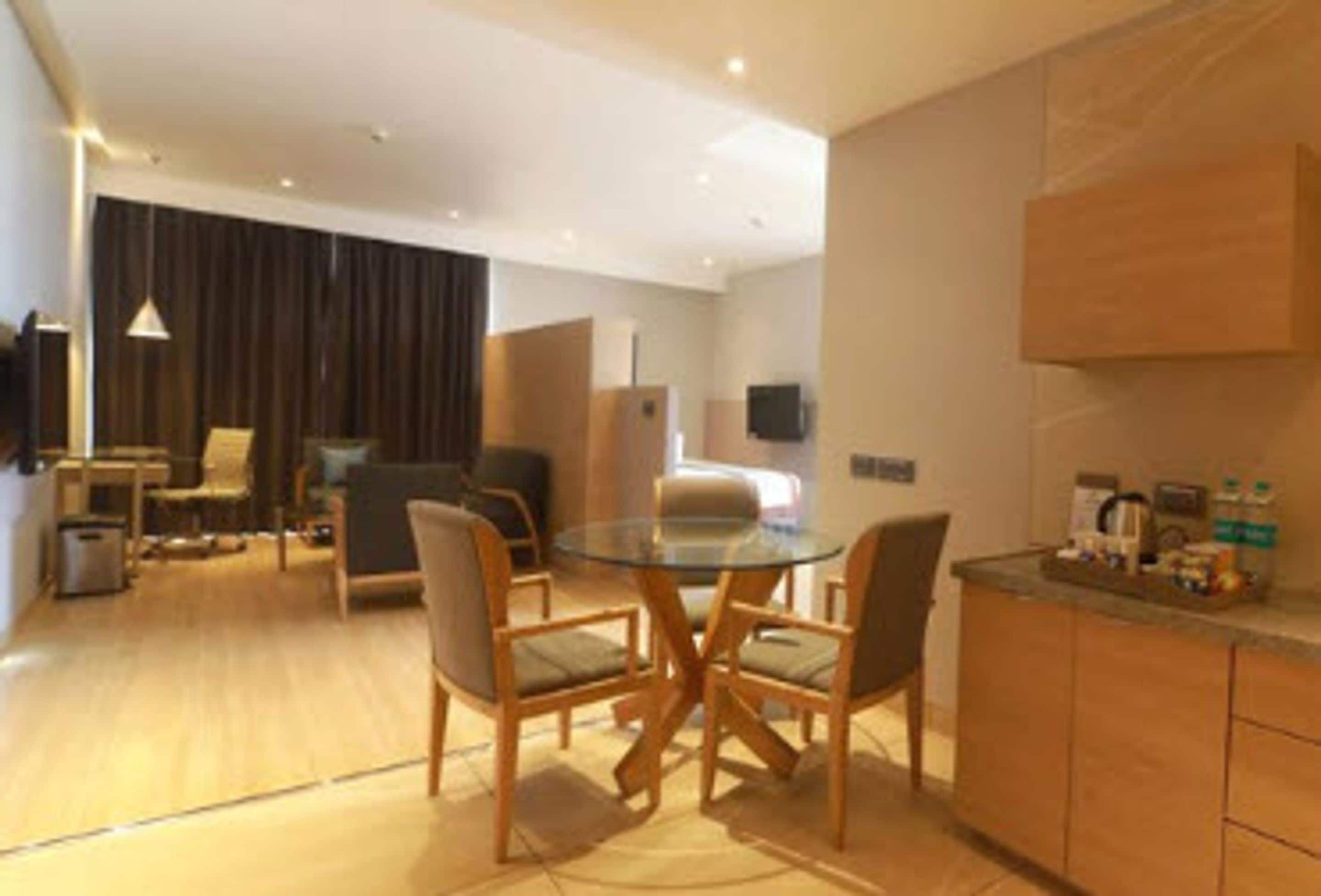 A modern hotel room with a glass dining table, wooden chairs, a sofa, a TV, and a kitchenette.