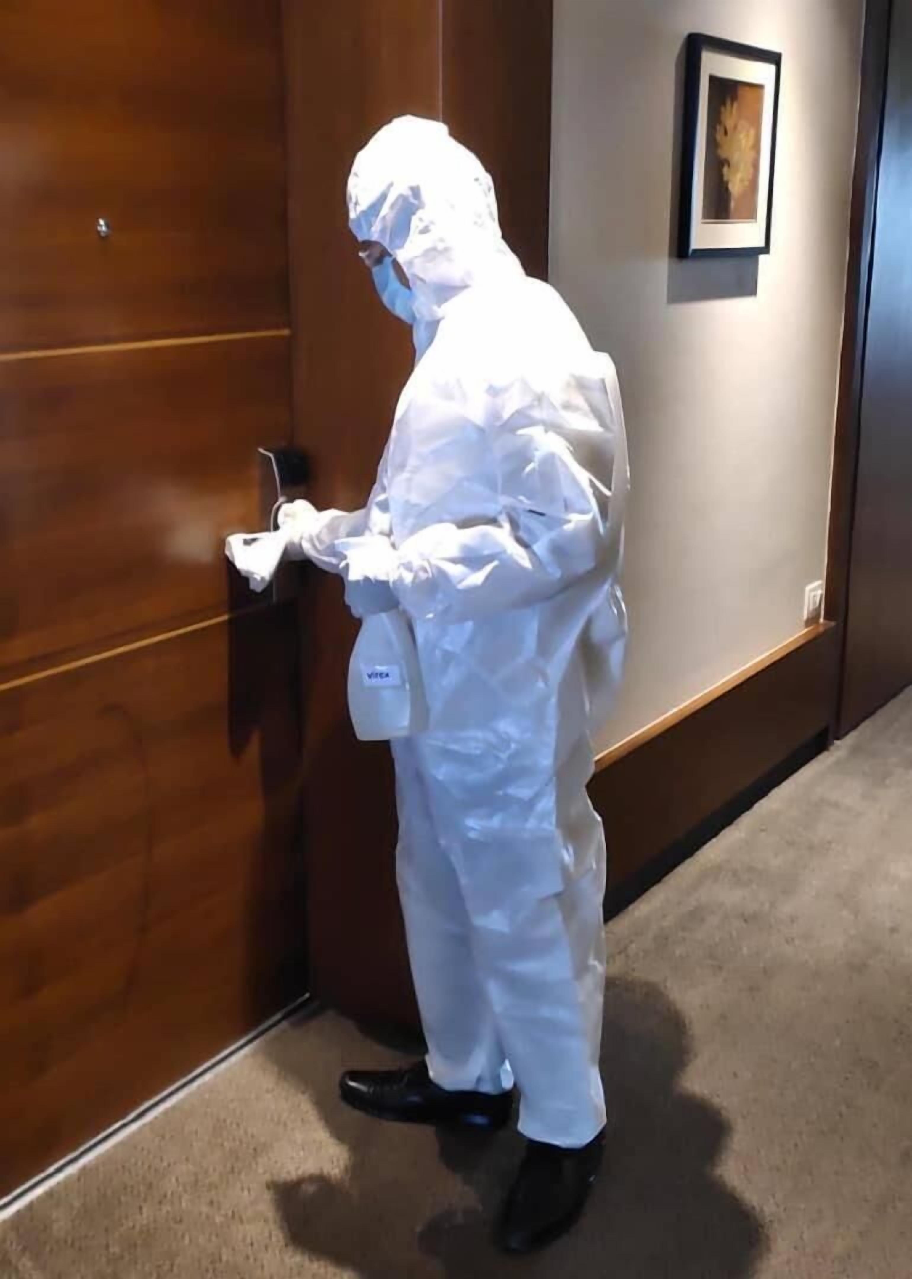 A person in a white protective suit standing in a hallway, holding a device near a door.
