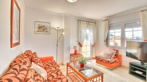 Un salon avec un canapé, un fauteuil et un téléviseur. Il y a des fenêtres avec des rideaux et une plante posée sur une table.