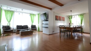 Una sala de estar amplia con techo de madera, cortinas verdes, un comedor con mesa y sillas de madera, y un televisor montado en la pared.