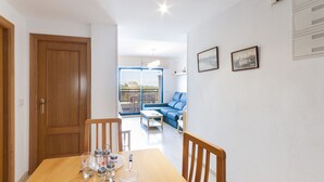 A dining area with a wooden table set for four, a blue sofa in the background, and a balcony with a view. - ALEJANDRITA - Apartment with shared pool in Playa De Gandia and Free Wifi (Gandia)