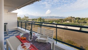 A balcony with a checkered floor, a table set with a bottle, glasses, and oranges, and a view of distant mountains and fields. - ALEJANDRITA - Apartment with shared pool in Playa De Gandia and Free Wifi (Gandia)