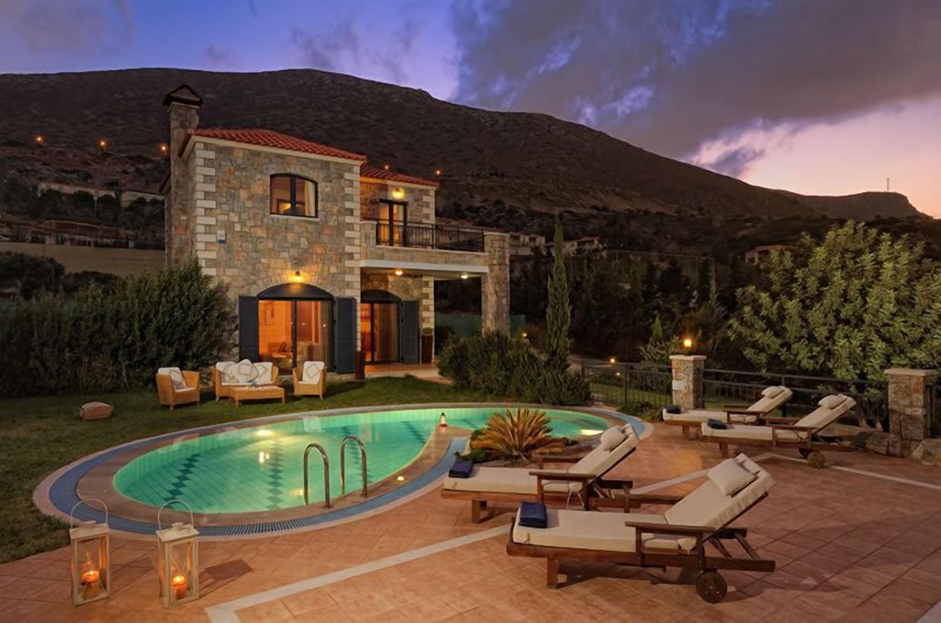 A two-story stone house with a swimming pool, outdoor seating, and a mountainous backdrop.