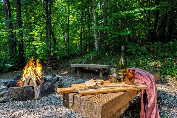 A campfire with a wooden table set with a bottle, glasses, and a blanket. - Perfect mountain cottage ☎ Fire pit ☎ Gas Fireplace ☎ Screened Porch ☎ Secluded (Townsend)