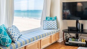 A room with a window seat overlooking a beach, a television on a stand, and a small table with electronic devices. - Ocean Dunes 1701 (Kure Beach)