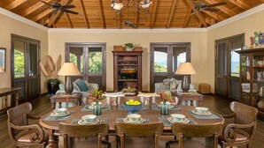A dining room with a large wooden table set for six, surrounded by six chairs. There are two windows with wooden shutters, a ceiling fan, and a chandelier. - Francis Bay Estate: Luxury Villa! Pool! Walk To The Beach! (Mary Point)