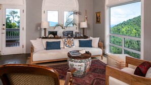 Ein Wohnzimmer mit Sofa, Couchtisch und Sesseln, einem Kamin und einem großen Fenster mit Blick auf einen Wald.
