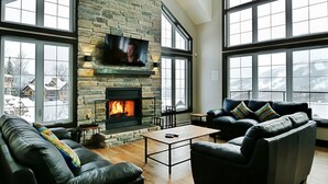 A spacious living room with a stone fireplace, leather sofas, and large windows offering a view of a snowy landscape. - Luxury cottage, panoramic view over the entire valley/Ski Resort, 24 people (Stoneham-et-Tewkesbury)