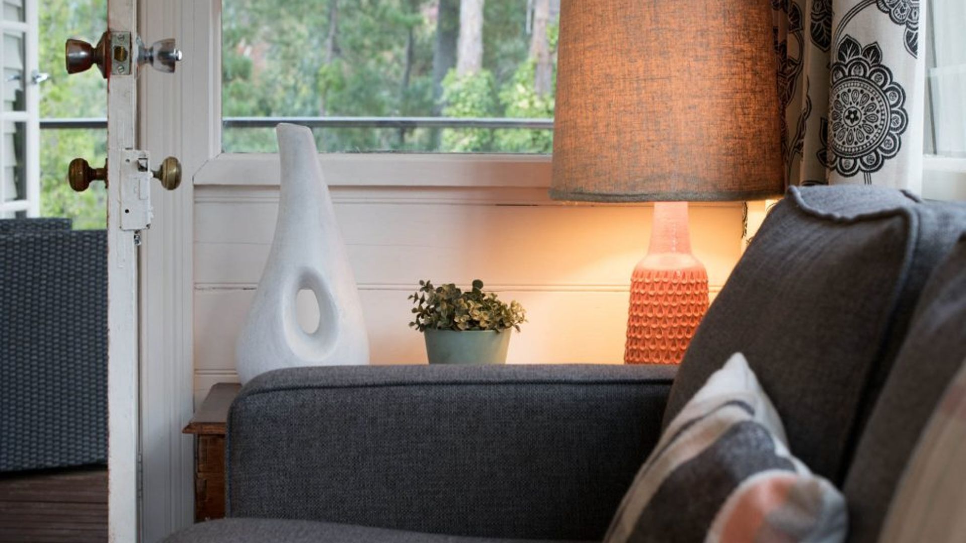 A room with a grey sofa, a potted plant, a lamp, and a window with a view of trees.