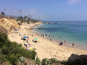 A beach with people sunbathing and swimming, surrounded by palm trees and a rocky cliff.